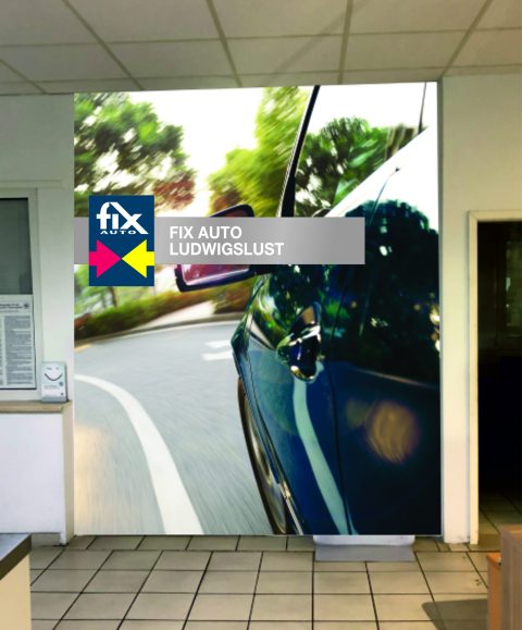 Ein Aufsteller auf einem Fliesenboden. Darauf ist das Bild eines auf der Straße fahrendem Auto, mit dem Fix Auto Logo in der oberen Mitte für Fix Auto Ludwigslust