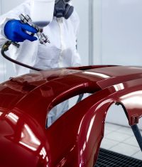 Car painter in protective clothes and mask painting automobile bumper with metallic paint and varnish in chamber workshop.