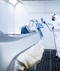 Car painter in protective clothes and mask painting automobile bumper with metallic paint and varnish in chamber workshop.