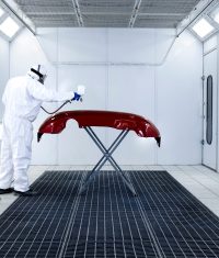 Car painter in protective clothes and mask painting automobile bumper with metallic paint and varnish in chamber workshop.