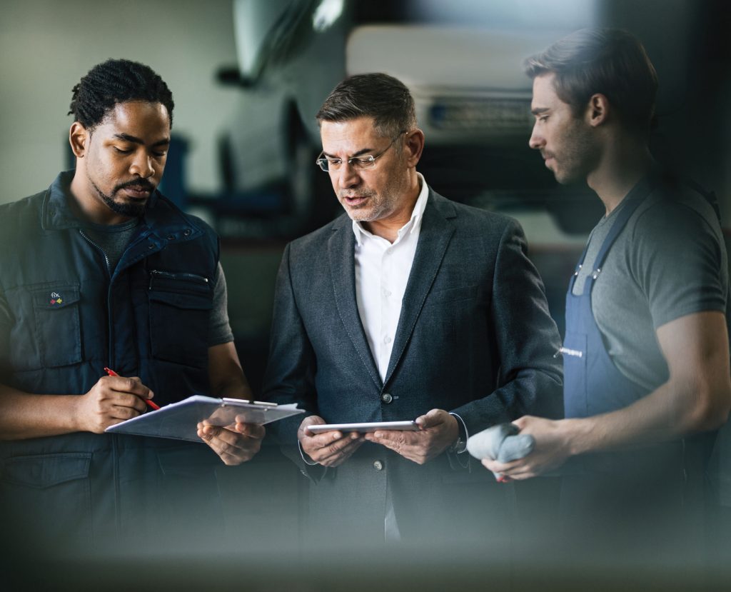 3 men talking, one with a clipboard, one with a tablet, and one with a shop rag