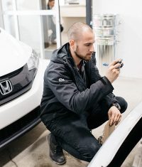Auto body technician inspecting vehicle damage and documenting details with a phone in a repair shop